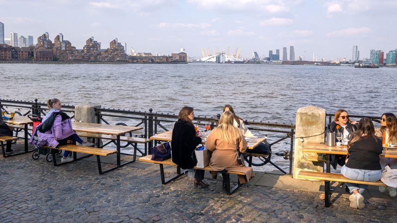 A photo of people sat on benches along the riverside at the Trafalgar Tavern with the river Thames and the City of London in the distance