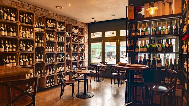 A wide shot of a wine bar with a wall lined with many bottles of wine and wooden tables and chairs in the centre.