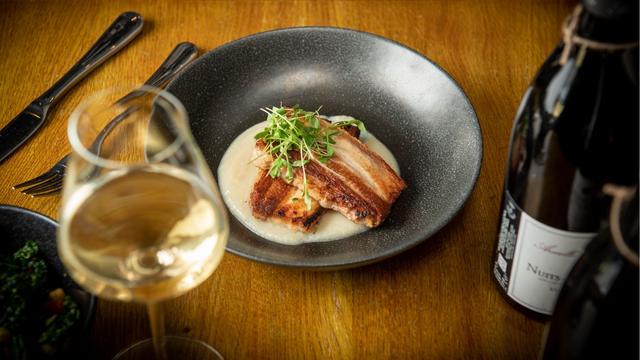 Pair expertly prepared dishes with delicious wines at Humble Grape wine bar. Image courtesy of Cru Comms. A table with a black plate with a fine dining dish of pork and sauce next to a bottle of wine and glass of white wine.