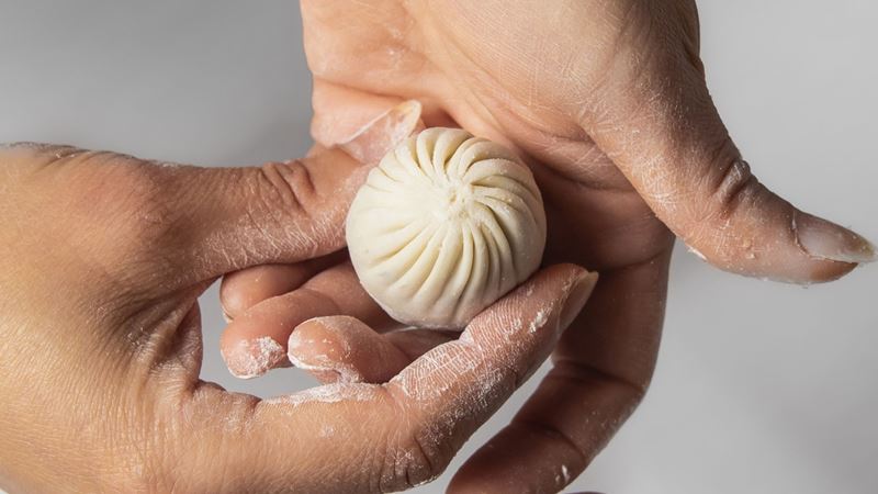 A photo of two hands covered in flour holding a Chinese dumpling that has just been rolled up