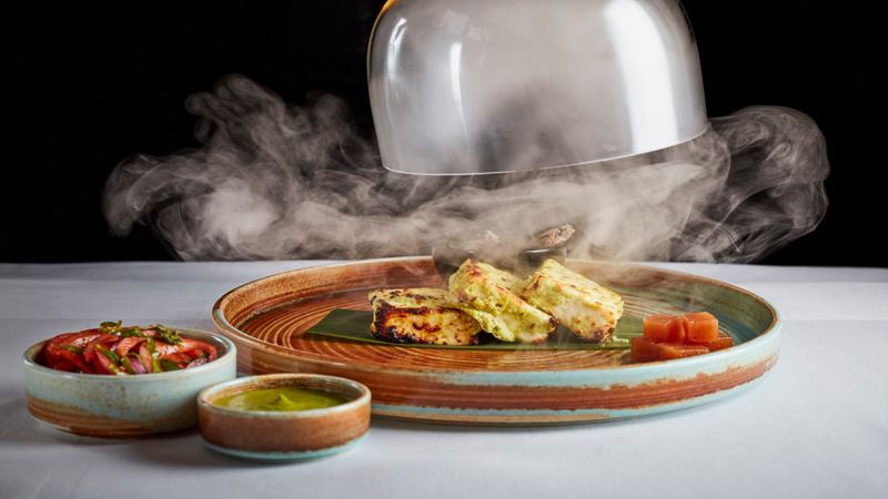 Savour signature Indian dishes with a luxurious flair at Benares in Mayfair, Credit: Jodi Hinds. Image courtesy of Benares. A cover is being lifted from a plate of food and steam billows out across the table.