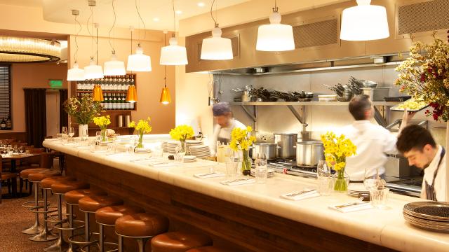 Grab a seat at the bar and watch the chefs at work. Image courtesy of Bocca Di Lupo. A white bar with brown stools and chefs cooking.