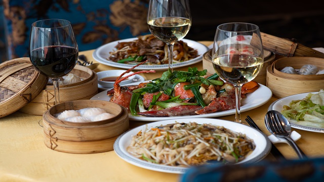 landscape shot of different plates of chinese food including noodles and dumplings, accompanied by glasses of red and white wine