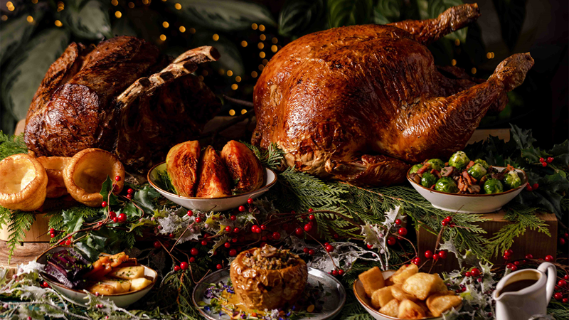A golden roast turkey sits on a table alongside bowls of crispy roast potatoes, cabbage, brussel sprouts, parsnips and yorkshire puddings.