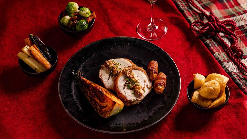 A traditional Christmas dinner at Straits Kitchen at Pan Pacific London. Credit: Pan Pacific London. Image courtesy of Pan Pacific London. A plate of stuffed turkey and pigs in blankets with small bowls of roast potatoes, roasted carrots and parsnips and brussel sprouts.
