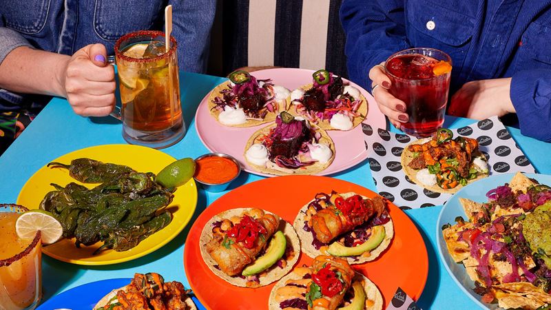 A photo of a table full of different colourful mexican dishes and drinks at Club Mexicana with two people's hands in shot holding glasses