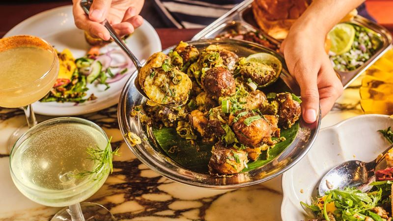 Choose from a variety of vibrant gluten free dishes at Dishoom. Credit: Dishoom. Image courtesy of Dishoom. A bird's eye photo of someone holding a dish of gunpowder potatoes at Dishoom with other dishes and cocktails on a table underneath