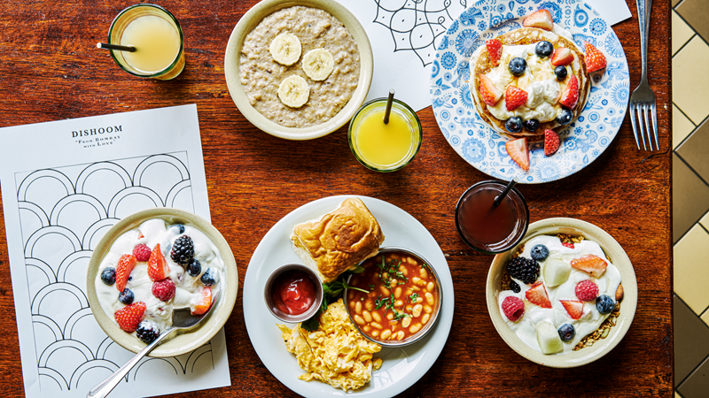 Keep kids and babies entertained with the colouring sheets at Dishoom while you enjoy a delicious brunch or dinner. Credit: Haraala Hamilton. Image courtesy of Dishoom. A bird's eye view of a wooden table with six plates of brunch plates and glasses of juice, including a savoury plate of scrambles eggs and beans, bowls of yoghurt with granola and fresh fruit, porridge and a pancake stack with kids colouring sheets underneath