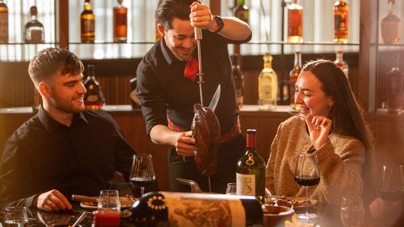Let the food keep flowing at Fazenda with its menu of prime meats and mouthwatering sides. Credit: Fazenda. Image courtesy of Fazenda. A photo of a couple sat at a table with a glass of wine each and a waiter carving a joint of meat at the table and serving for them