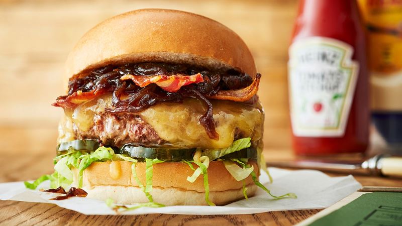 Take your pick from a menu of juicy options at Honest Burgers. Credit: Honest Burgers. Image courtesy of Honest Burgers. A close up photo of a burger at Honest Burgers with a juicy beef patty, melted cheese, salad and crispy bacon with a bottle of tomato ketchup in the background