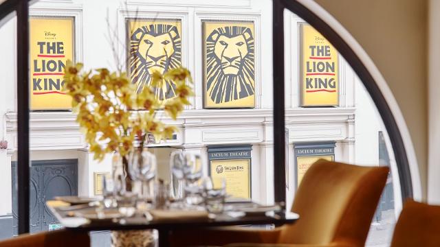 A brown table with a red chair in the window of indigo restaurant where diners can see the yellow posters of the lion king theatre show. 