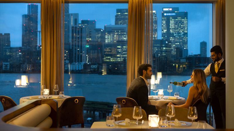 Indian fine dining with a view. Credit: InterContinental London – The O2. Image courtesy of InterContinental London – The O2. A cosy restaurant with ambient lighting and a couple sat in front of the wall of windows with a view over the London city skyline at dusk.