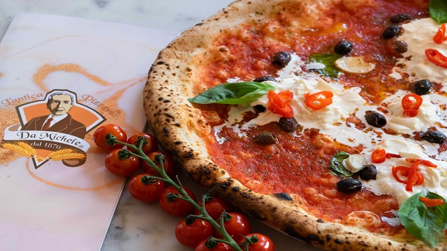 Pizza at L'Antica Pizzeria da Michele. Image courtesy of L'Antica Pizzeria da Michele. Fresh pizza topped with cheese, olives and basil leaves, juxtaposed with a vine of tomatoes and a napkin