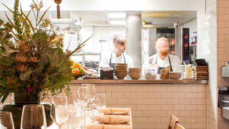 Browse the menu of Mediterranean delights at NOPI in Soho. Credit: NOPI. Image courtesy of Ottolenghi. A photo of two chefs in the kitchen from the dining area with stacks of bowls and plates on the counter and a table covered in crockery, glassware, cutlery and flowers in the foreground