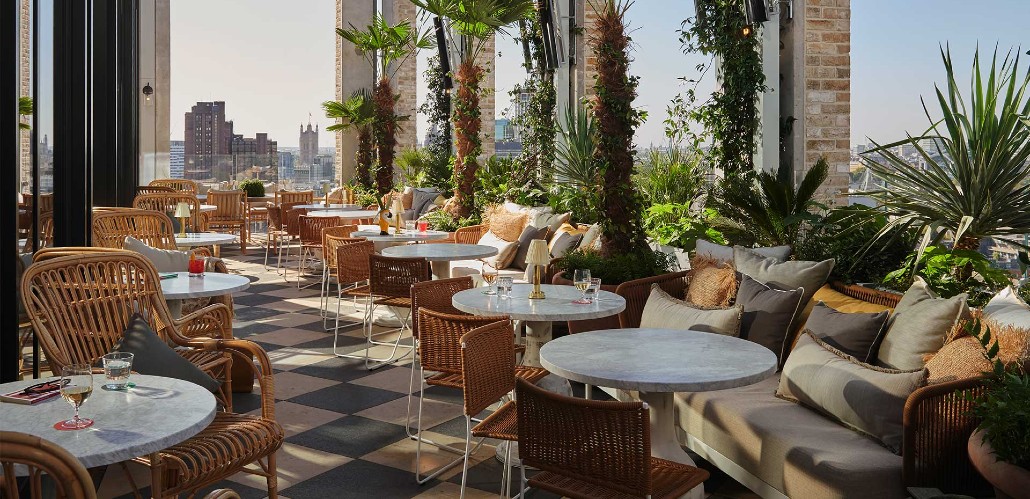 Rooftop terrace restaurant with chairs and tables set out or dining, with London skyline in distance.