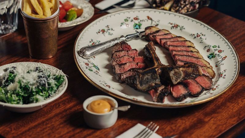 Steak is served on the bone for extra flavour at cult-favourite, Blacklock. Credit: Al Ward/Pine Studios. Image courtesy of Blacklock. A photo of a vintage dish with a perfectly cooked T bone steak thinly sliced on top, with sides of fries and salad on a brown wood table at Blacklock