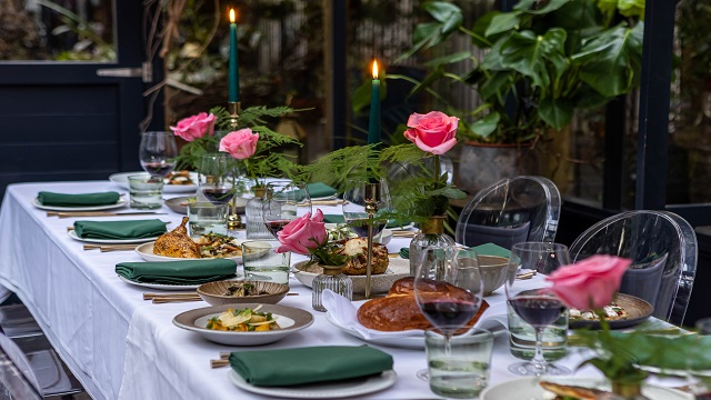 a long table decorated with a white tble cloth, green napkins and glasses, pink roses down the middle of the table and greenery in the background