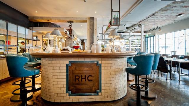 Perch at the counter of this delightful modern cafe. Image courtesy of The Riding House Cafe. A beige restaurant with a round dining counter and large blue stools.