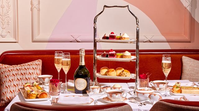 Afternoon tea trays with cakes and scones, plates with finger sandwiches, a bottle and two glasses of champagne, plus tea, laid out on white tablecloth with red banquette seating and cushions in the background.