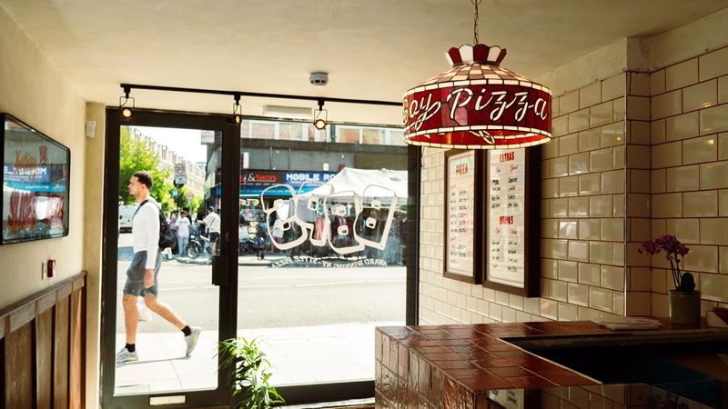 Enjoy a slice or three at Bad Boy Pizza Society spots across London. Credit: Josh Oliver/Bad Boy Pizza Society. Image courtesy of Bad Boy Pizza Society. A photo of the interior of the Bad Boy Pizza Society shop in Bethnal Green with white tiles, a personalised mirror and glass lampshade with red tile counter