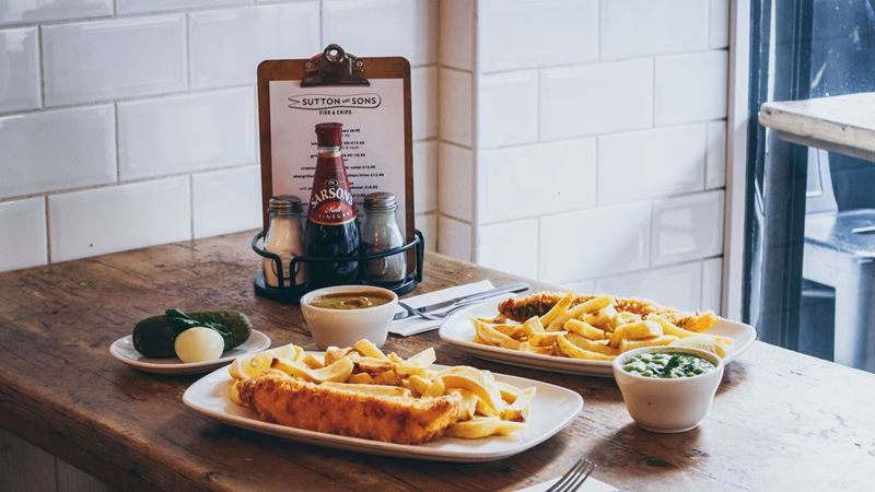 A photo of two plates of fish and chips on a wooden table at Sutton and Sons with condiments and a menu on the table