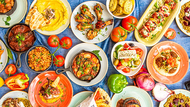 Tuck into Greek and Mediterranean cuisine © The Real Greek. Image courtesy of The Real Greek. A blue table spread covered with tomatoes and peppers and plates of colourful Greek and Mediterranean food