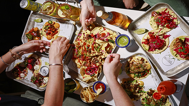 A group of friends are sharing a selection of tacos and nachos disposed on trays at Club Mexicana.