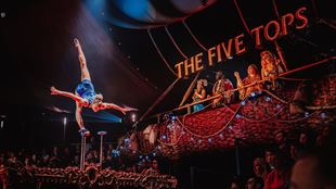 circus tent with an audience watching an acrobat do the splits on an elevated stage in a spotlight