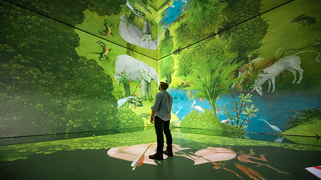 A man stands in one of the galleries at FRAMELESS and observes the painting The Garden of Earthly Delights projected on two walls.