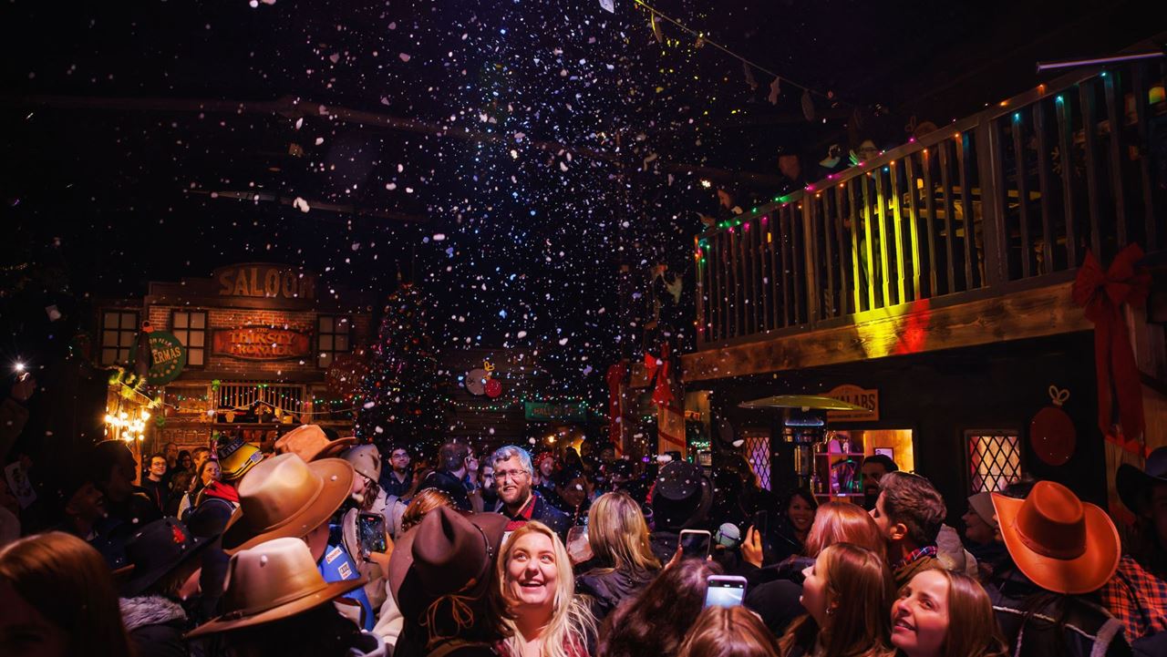 Celebrate Wintermas at Phantom Peak with festive fun, unique puzzles and deliciously festive food and drink! © Phantom Peak/Alistair Veryard Photography. large crowed wild west theme town with fake snow falling from the ceiling and groups of people looking up and smiling