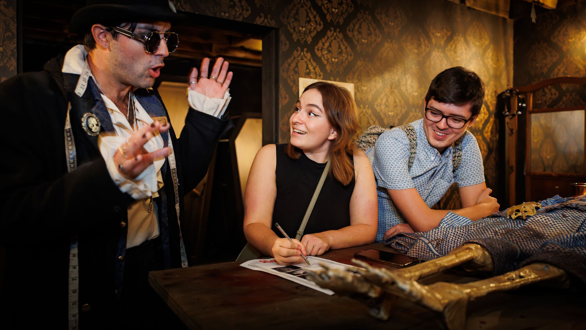Meet quirky characters and take part in exciting immersive missions. © Golden Tours/Alistair Veryard Photography. an actor wearing a long dark dark and black sunglasses, gesturing to two people smiling and taking notes