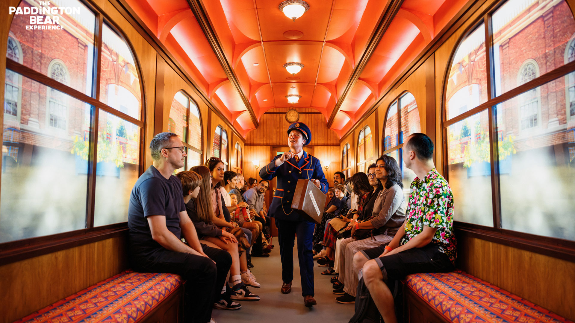 Start your adventure at Paddington Bear Experience with the Station Master and hop on a carriage for a ride into the city. Image courtesy of Golden Tours. Visitors are seated in a large vintage railway carriage as the Railmaster walks by.