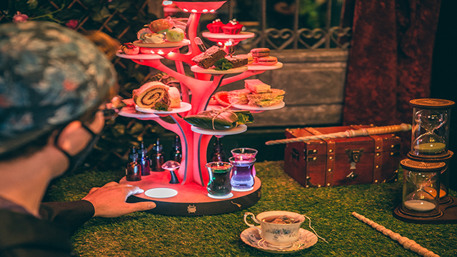 Wizard Afternoon Tea at The Wands and Wizard Exploratorium. Image courtesy of The Cauldron A man touches the afternoon tea stand in form of an pink illuminated tree where are disposed a variety of cakes and a selection of molecular drinks.