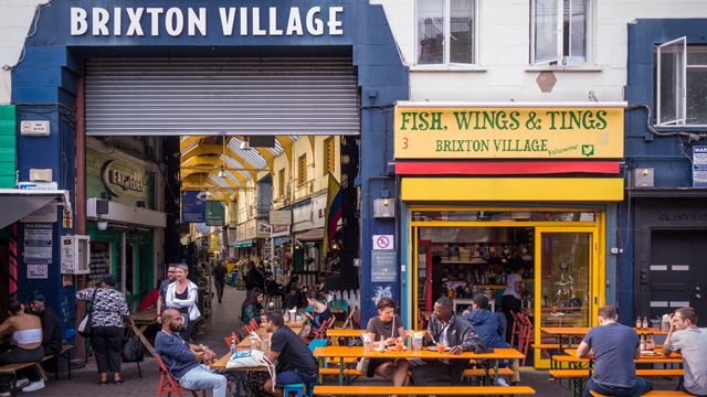 Entrance framed by dark blue sign to the covered arcade of Brixton Village, with Fish, Wings and Tings yellow-fronted restaurant next to it.