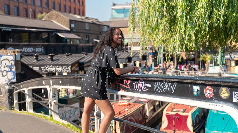 A person stands on a bridge and smiles while looking across Camden Lock