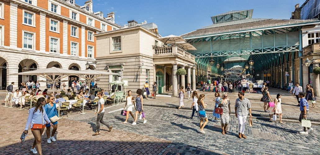 Shoppers walk around Covent Garden's West Piazza, in front of the market building, on a bright, cloudless day.