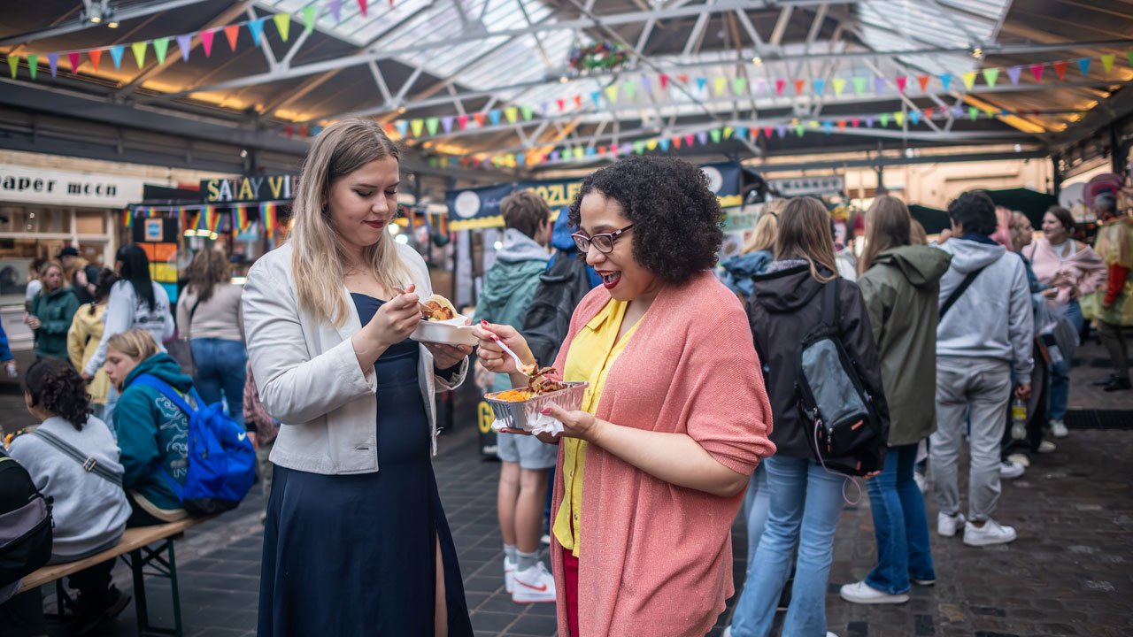 Enjoy a bit of shopping at Greenwich market before treating yourself to some delicious street food. © London & Partners/Michael Barrow Two women are standing in Greenwich market food court and sampling some of the street food sold there.