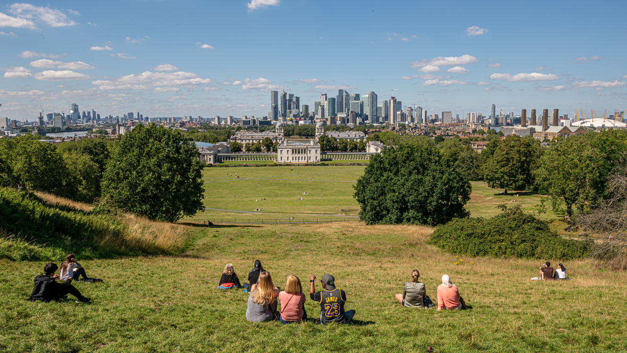 Skyline views from Greenwich Park. Image courtesy of Michael Barrow/London & Partners. A few people are sitting on top of the hill overlooking Greenwich Park and Canary Wharf.
