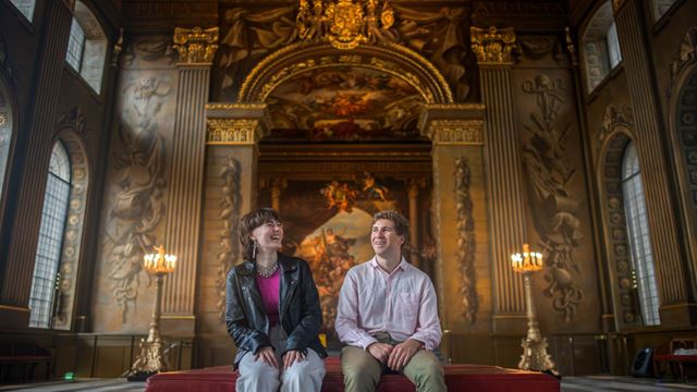 Two people sit on a red bench looking up at the painted ceilings of the painted hall in greenwich.