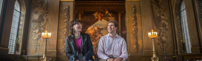 Two people sit on a red bench looking up at the painted ceilings of the painted hall in greenwich.