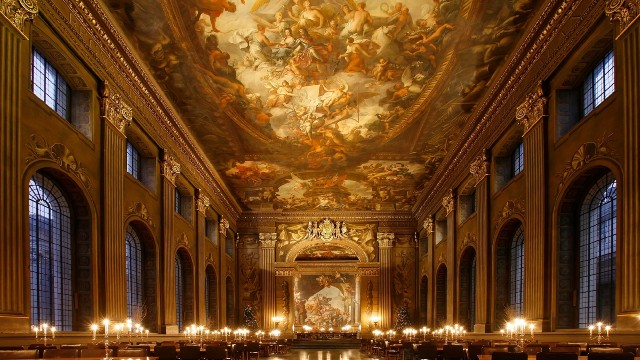 A highly ornate painted ceiling overhangs a large historic room lit by candles. 