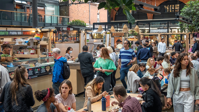 People gathered in Old Spitalfields Market buying food from stalls and eating it on nearby tables