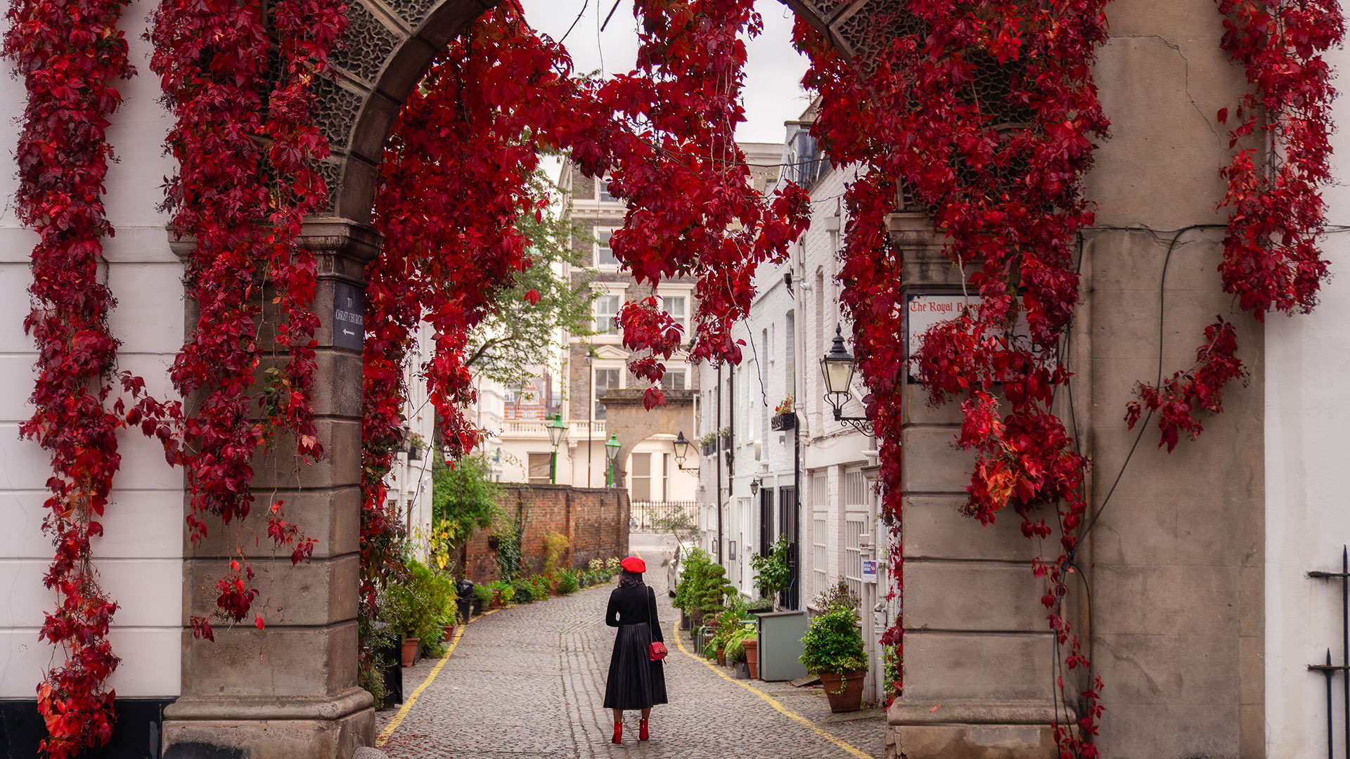 Seek out Kensington's colourful mews including its most famous, Kynance Mews. Credit: Antoine Buchet. Image courtesy of visitlondon.com. A woman dressed in a black coat and a red hat standing in front of Kynance Mews in Kensington, with red flowers hanging from the archway.