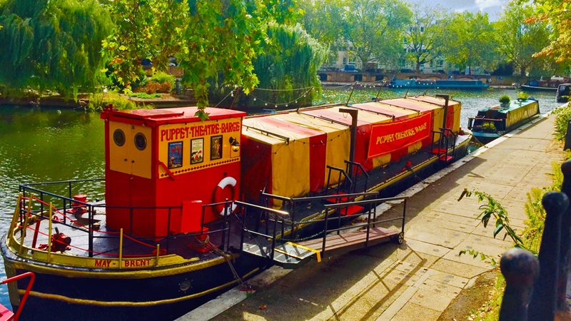 Entertain the whole family with a visit to the Puppet Theatre Barge in Paddington. Credit: Stan Middleton. Image courtesy of Puppet Theatre Barge. Red and orange barge on the river on a bright sunny day with Puppet Theatre Barge painted on the side