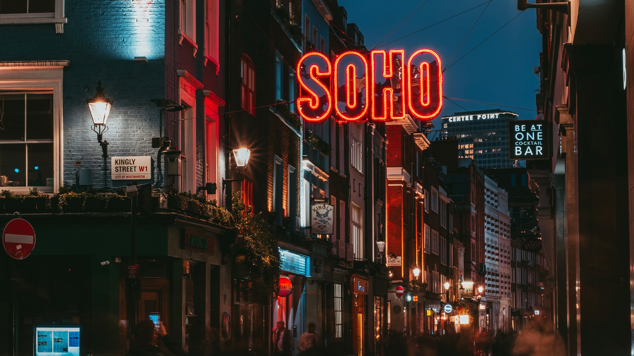 Discover the buzzing Soho area at night in London. Image courtesy of Sister London. A Soho street at night with a large neon sign saying Soho at the top of the picture.