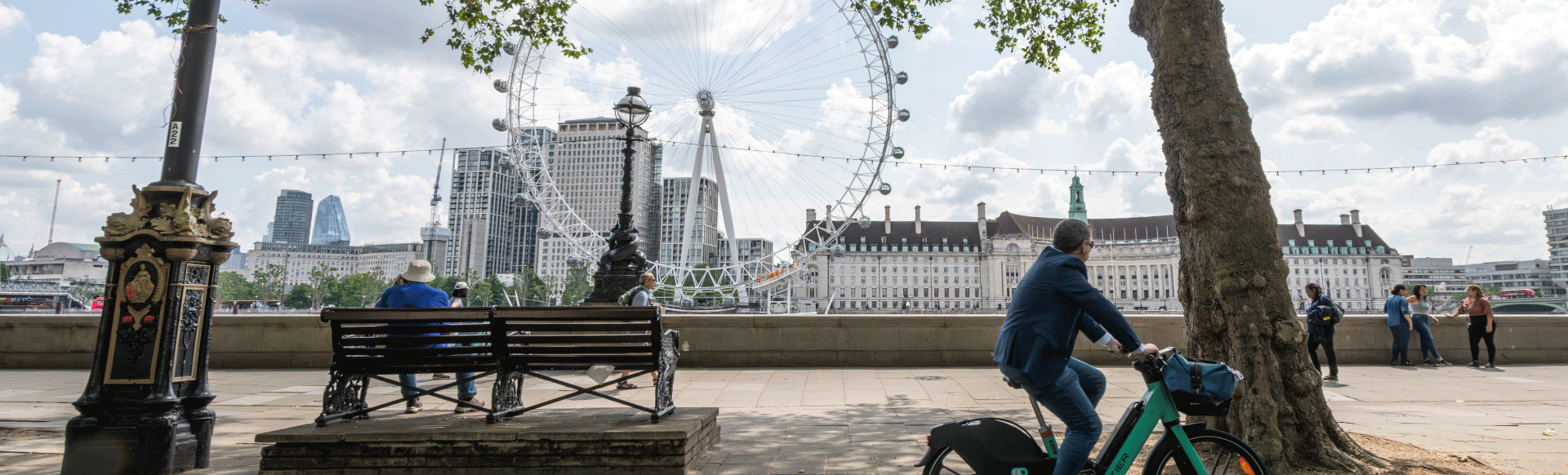 A man cycling a bike and another person sat on a bench at Westminster with the London Eye in the background