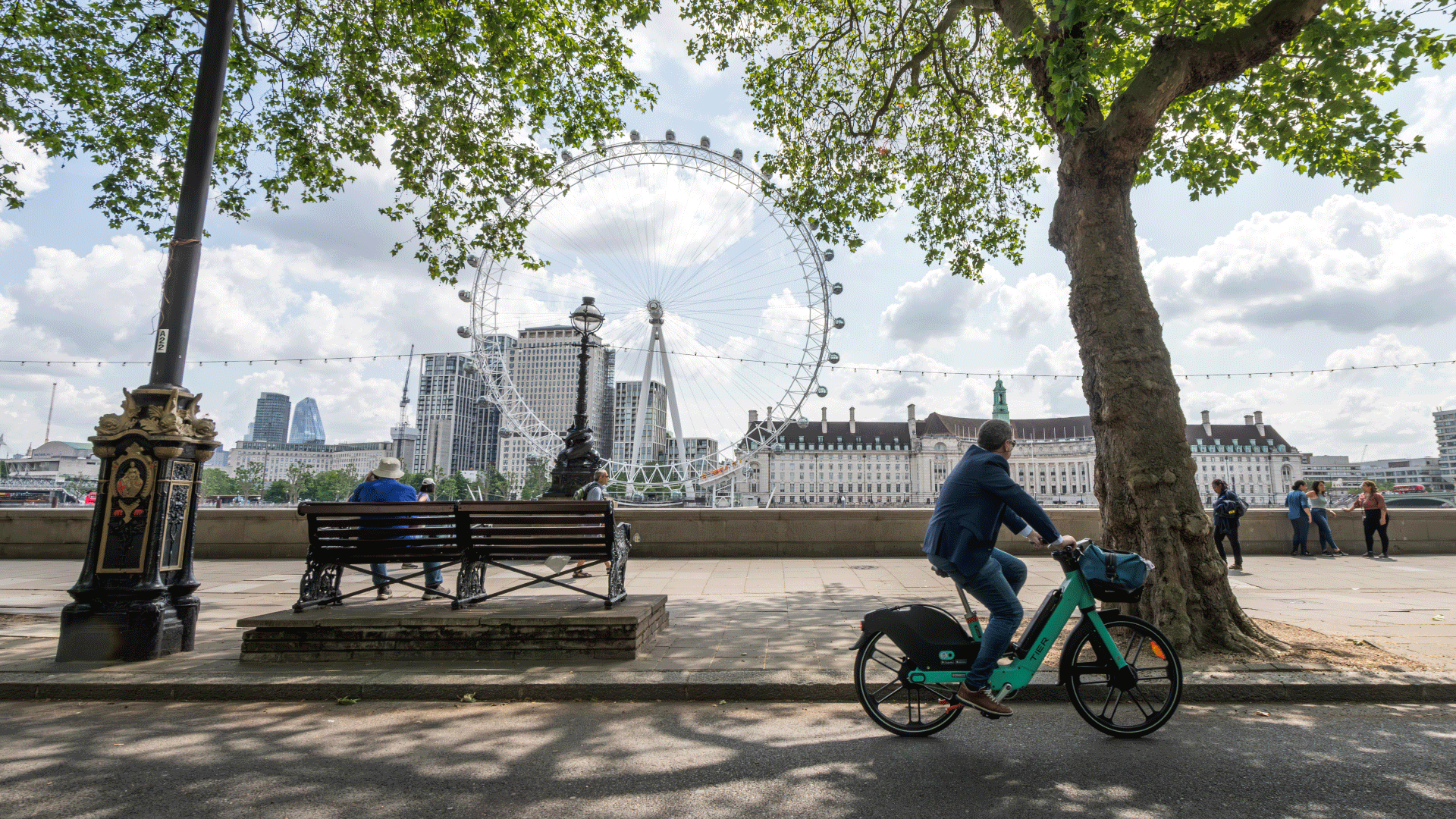 A view of the London Eye and Southbank from Westminster. Image courtesy of London & Partners/Michael Barrow. A man cycling a bike and another person sat on a bench at Westminster with the London Eye in the background