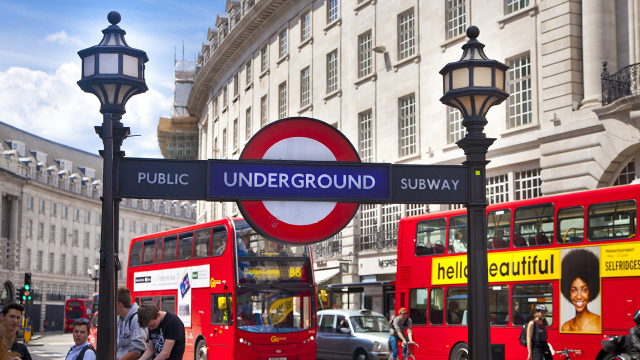 Piccadilly Circus London Underground sign with red buses and taxis driving past in the background.