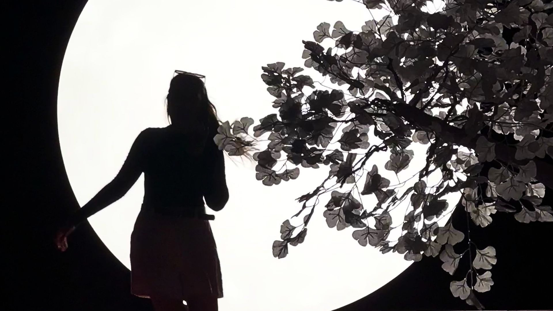 Capture your silhouette in the light of the moon at Moco Museum London. © Visit London/Kirstine Spicer A silhouette on a long haired woman in the light of a moon next to a tree.