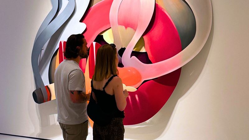 Two people stand in a tshirt and jeans in front of a large pair of lips smoking at moco museum.